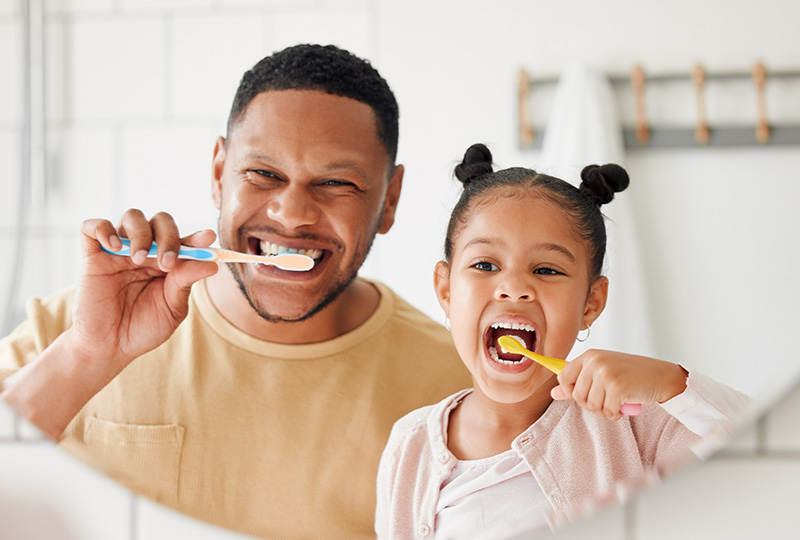 A man and a child are brushing their teeth together in front of a bathroom mirror.